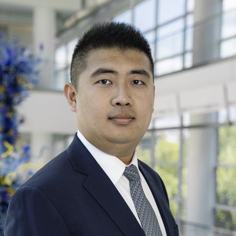 Professional headshot of an individual photographed from the chest up in an office or atrium setting. The individual is wearing a dark blue suit jacket, white dress shirt, and patterned tie, with large windows and architectural elements softly blurred in the background. A blue glass art installation is visible along the left side of the image, and natural light illuminates the indoor space.