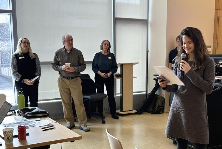 "A group of people stand near large windows in a bright indoor space, with one person holding a microphone and reading from a sheet of paper. Three individuals stand together near a lectern, some holding notes or drinks. In the foreground, a table holds laptops, papers, and a drink cup. The room has light-colored floors and tall window shades that diffuse the natural light."